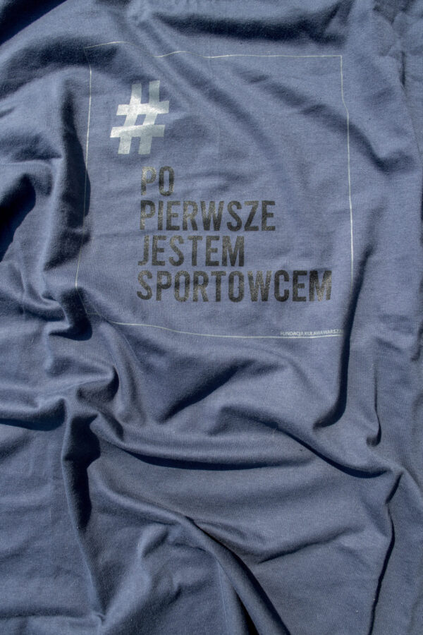 grafitowo niebieska koszulka z logo kampanii - prostokątna jasnoszara ramka z napisze # (w kolorze szarym) Po Pierwsze Jestem Sportowcem (w kolorze czarnym) małymi szarymi literami napis Fundacja Kulawa Warszawa