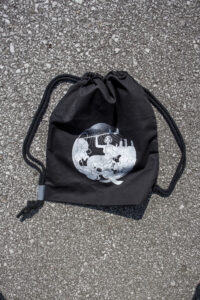 Czarny plecak w stylu worek, na grubych sznurkach z małymi kawałkami szarek odblasku u dołu. Nadruk na plecaku przedstawia syrenkę na wózku z kulą ręce
